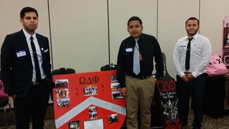 Three fraternity members wearing neckties posing next to their chapter display board