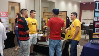 Two fraternity members in their chapter shirts talking with two attendees in front of display board