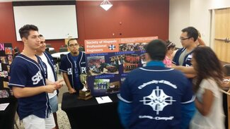 Five fraternity members in their chapter apparel talking with student in front of display board